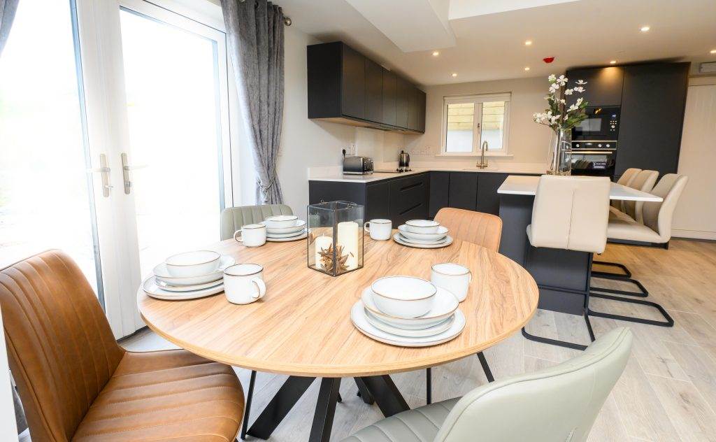 Modern kitchen and dining area with a wooden table set for six, featuring sleek black cabinets and c.