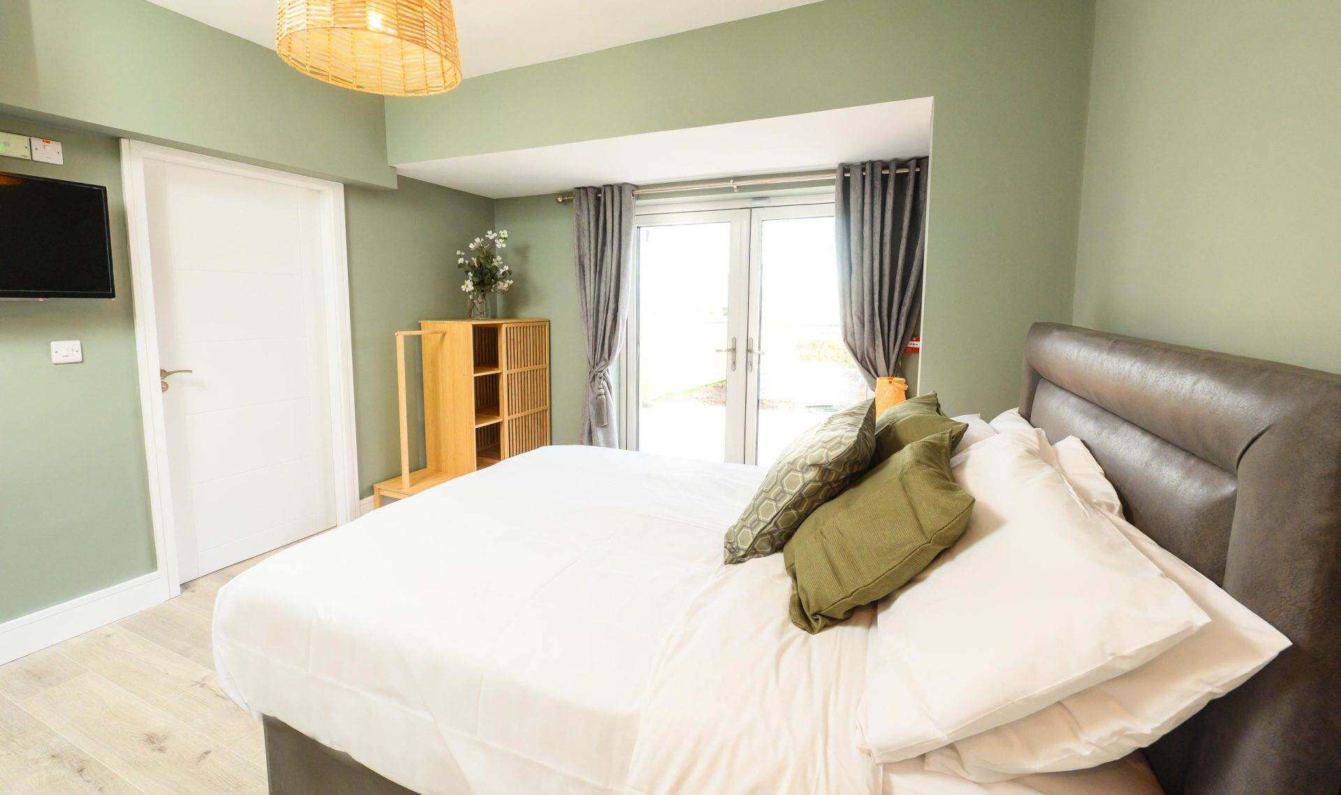 Modern bedroom with large bed, green walls, and natural light from balcony doors.