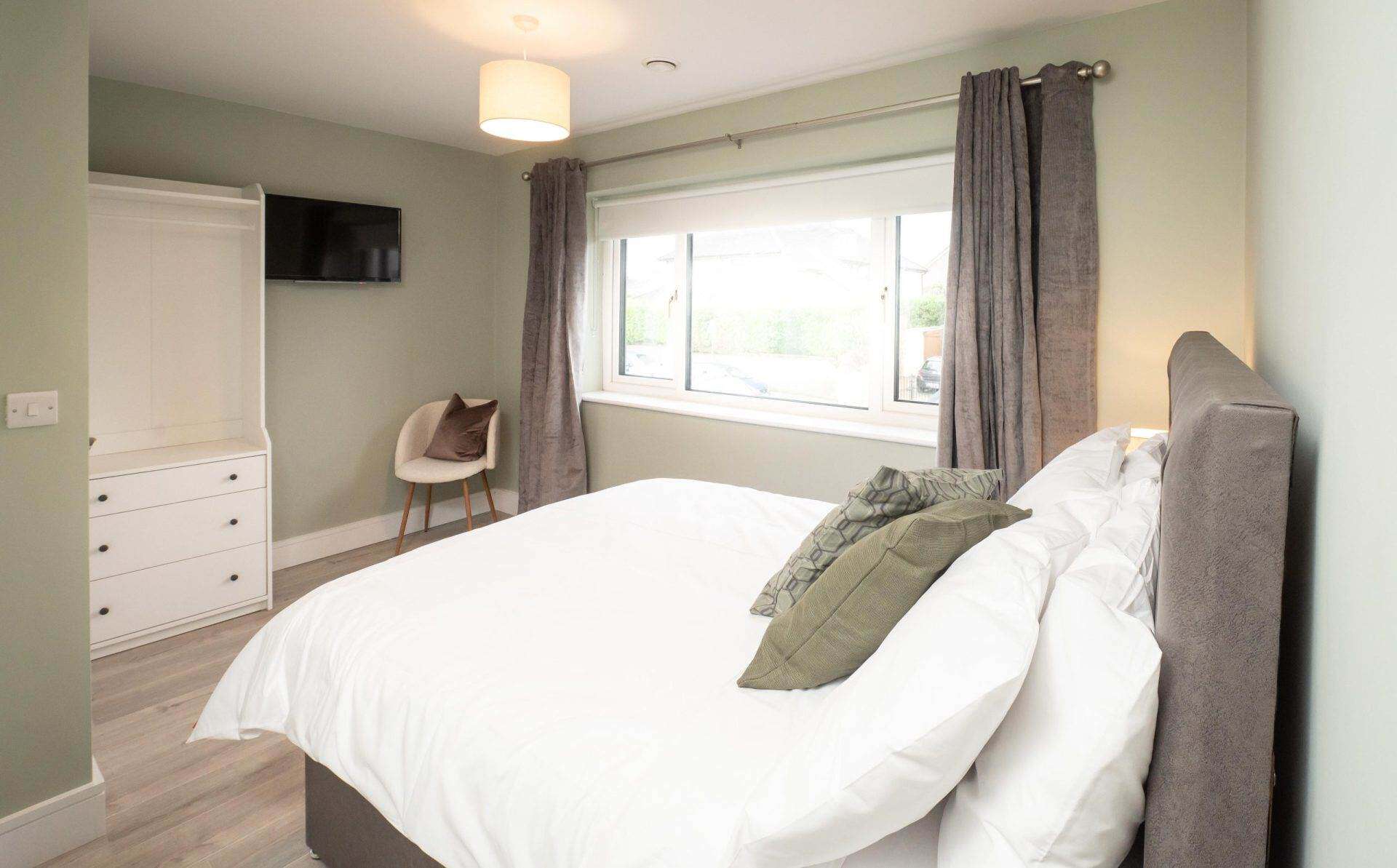 A comfortable bedroom with a large window, grey curtains, and a neatly made bed with pillows.